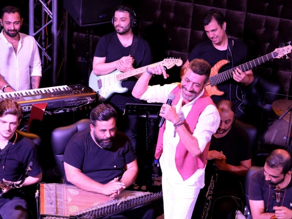 Hakan Altun elinde mikrofon tutarken iki gitarist ve diğer çalgıcılarla mor ışık altında sahneyi paylaşıyor